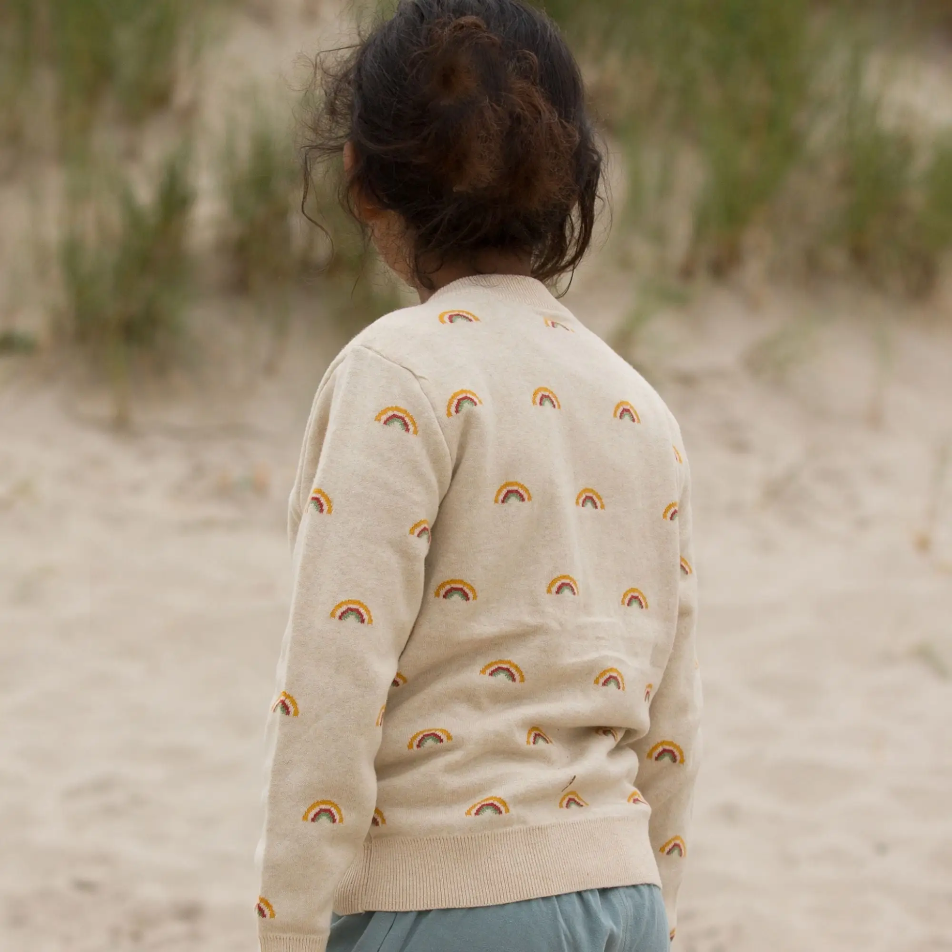 Child turned away from the cmera looking at the grasses at the beach From One To Another Rainbows Knitted Cardigan Sweater